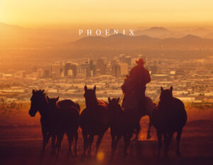 Welcome to Arizona: cowboy herds horses at sunset above the Phoenix skyline with layered desert mountains.